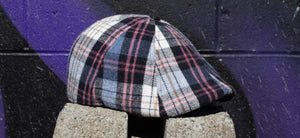 The Good Egg Cap, a plaid cap featuring light blue and pink sitting on a cement block in front of a colorful wall