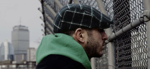 The Eire Scally Cap featuring a green plaid shown on a man looking through a chainlink fence